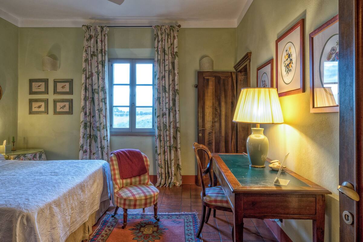 A cozy room with a bed, a chair, a desk with a lamp, and framed pictures on the wall.