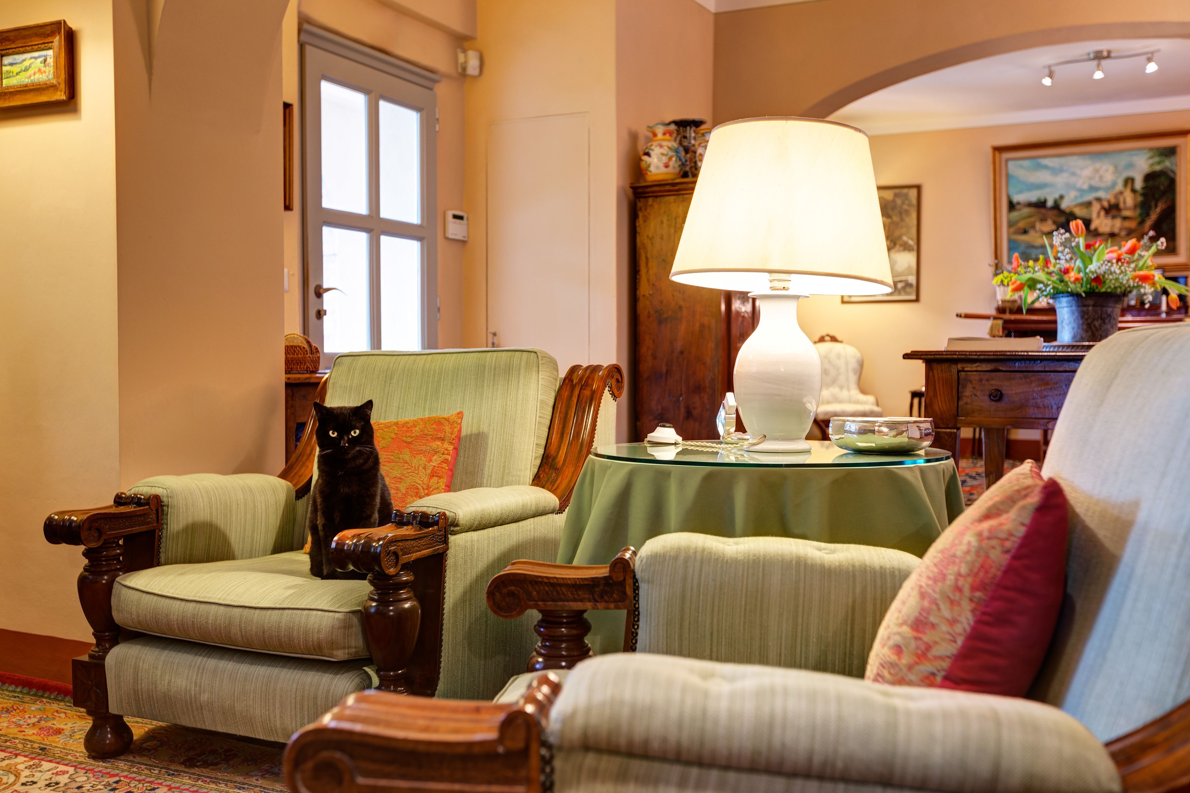 A cozy room with two upholstered chairs, a wooden table with a lamp, and a cat sitting on one of the chairs.