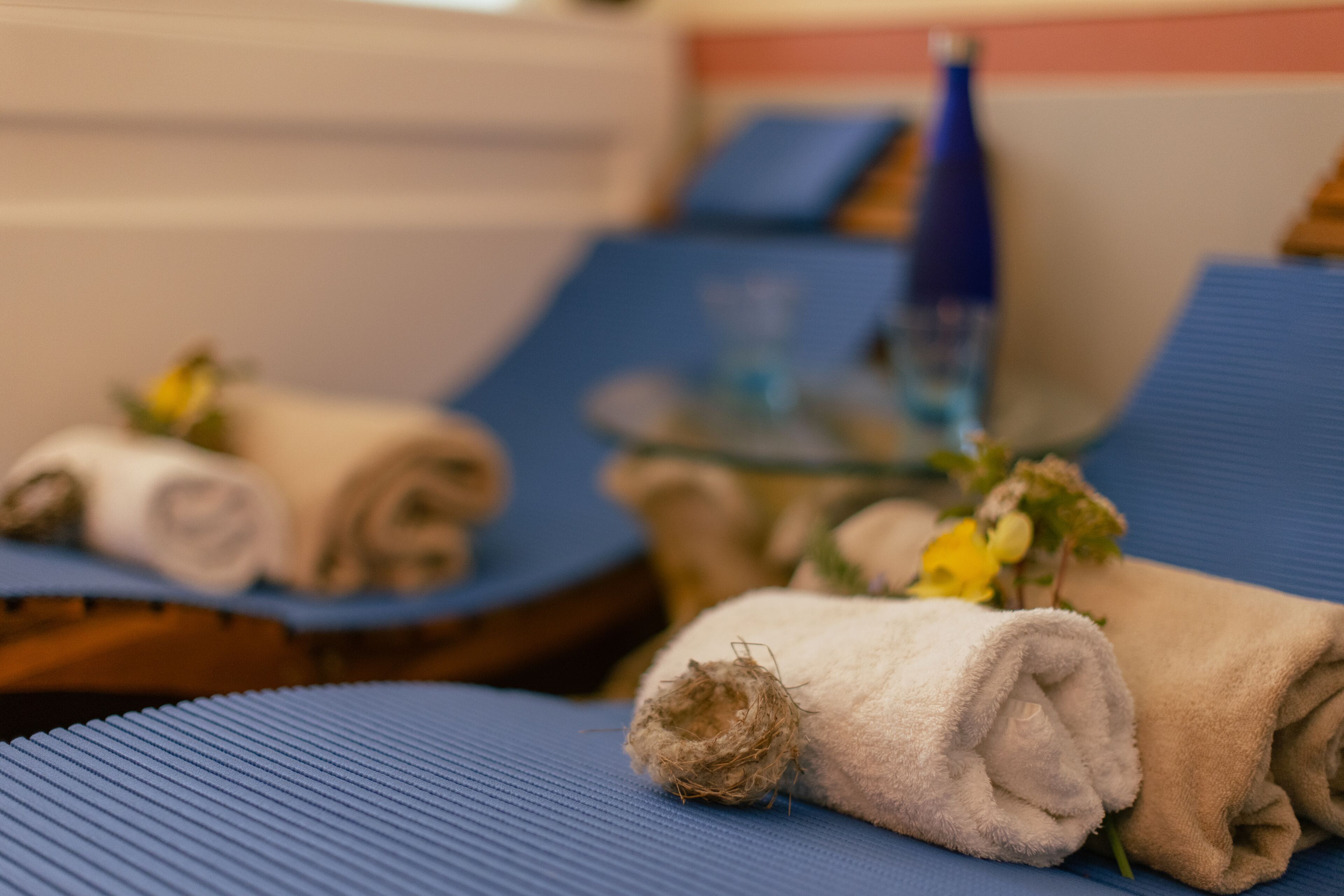 A room with blue cushioned chairs, neatly folded towels, and a glass table with a bottle and flowers.