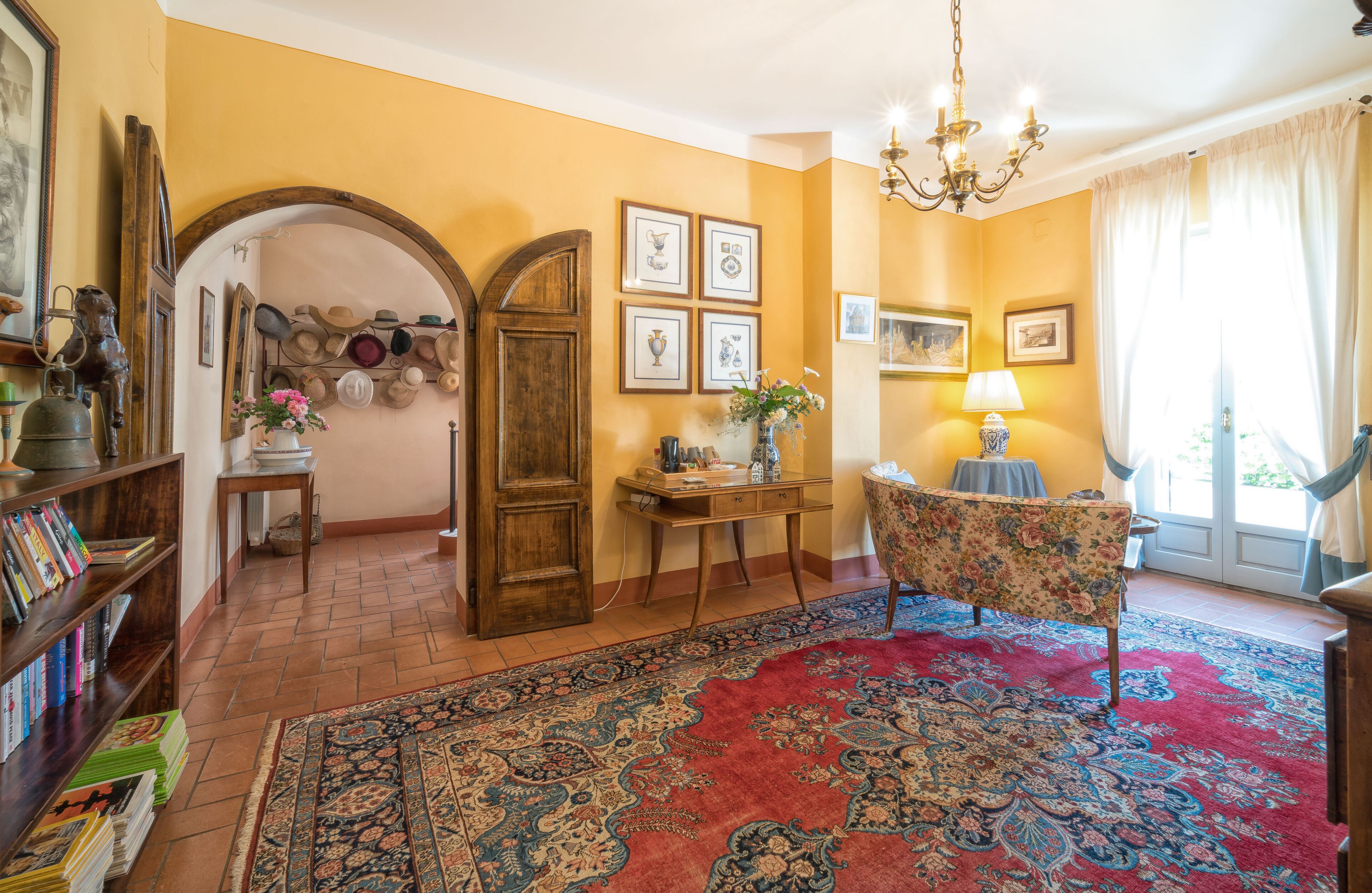 Una habitación con puerta de madera, una estantería, una mesa con un florero, una silla y una alfombra grande.