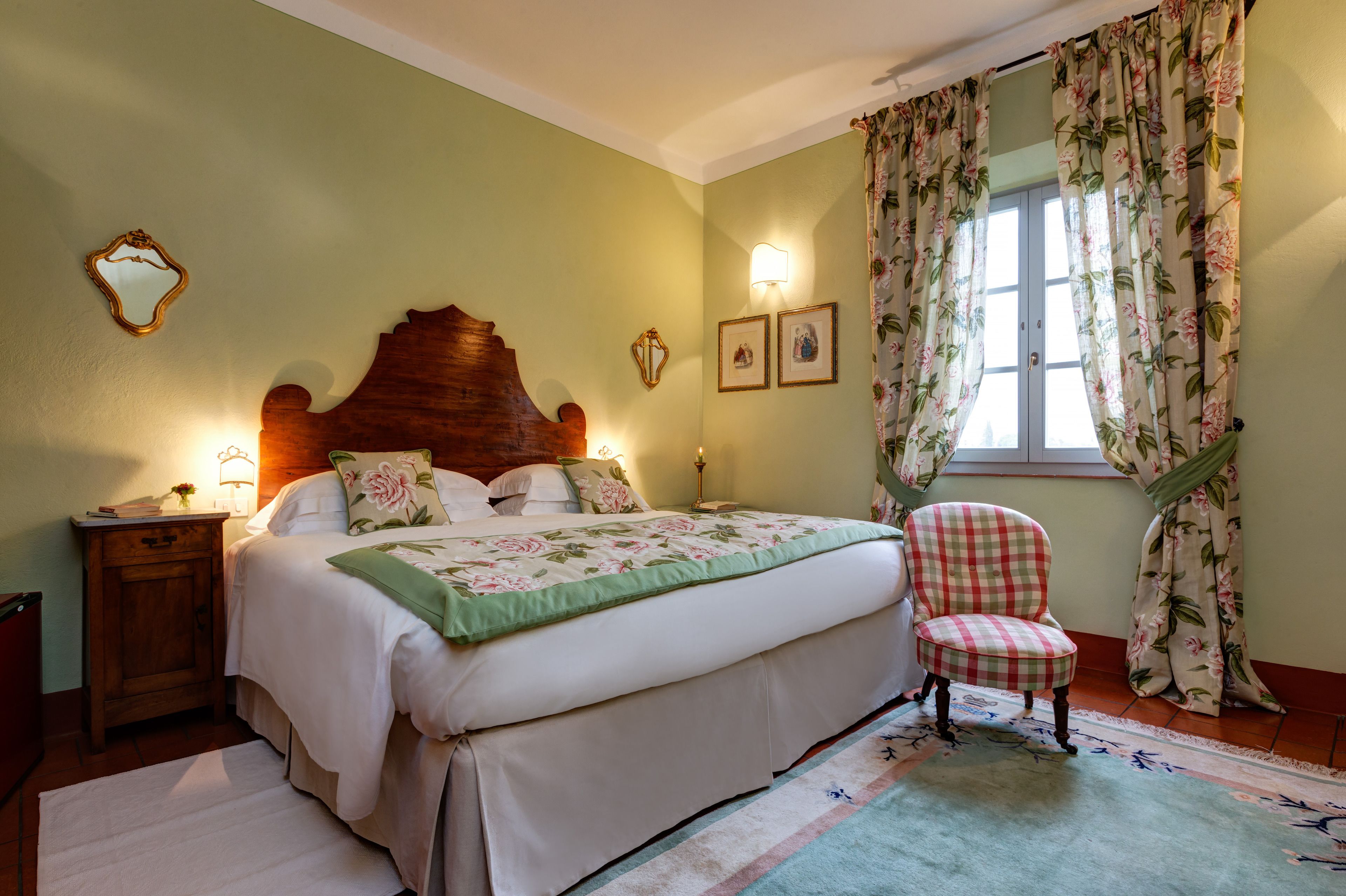 Un dormitorio con cama, una silla, una mesita de noche y una ventana con cortinas florales.