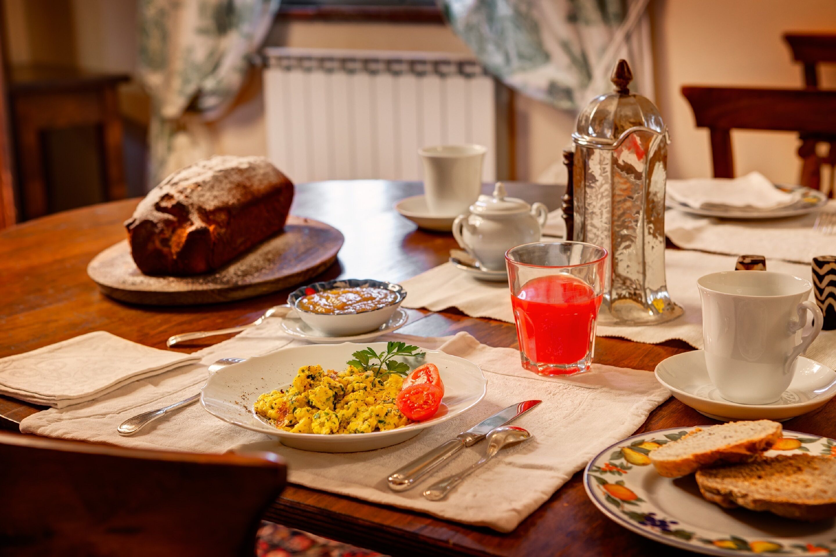 A breakfast setting with a loaf of bread, a plate of scrambled eggs, a glass of orange juice, and a teapot on a wooden table.