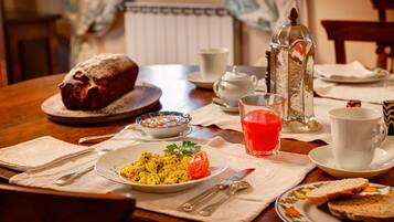 A breakfast setting with a loaf of bread, a plate of scrambled eggs, a glass of orange juice, and a teapot on a wooden table.