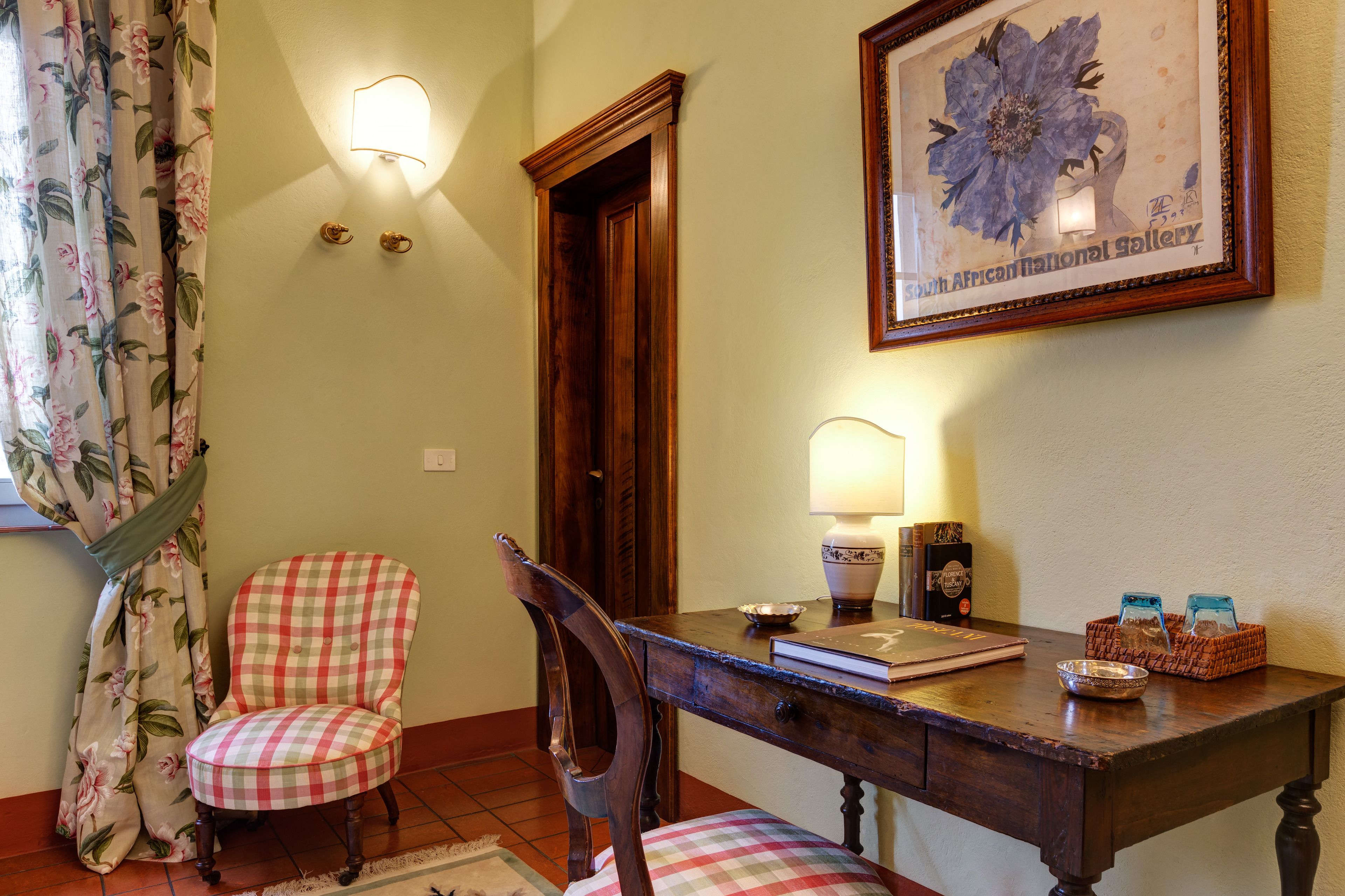 Un rincón comedor con mesa de madera, dos sillas, una lámpara, una obra de arte enmarco y un cortinaje floral.