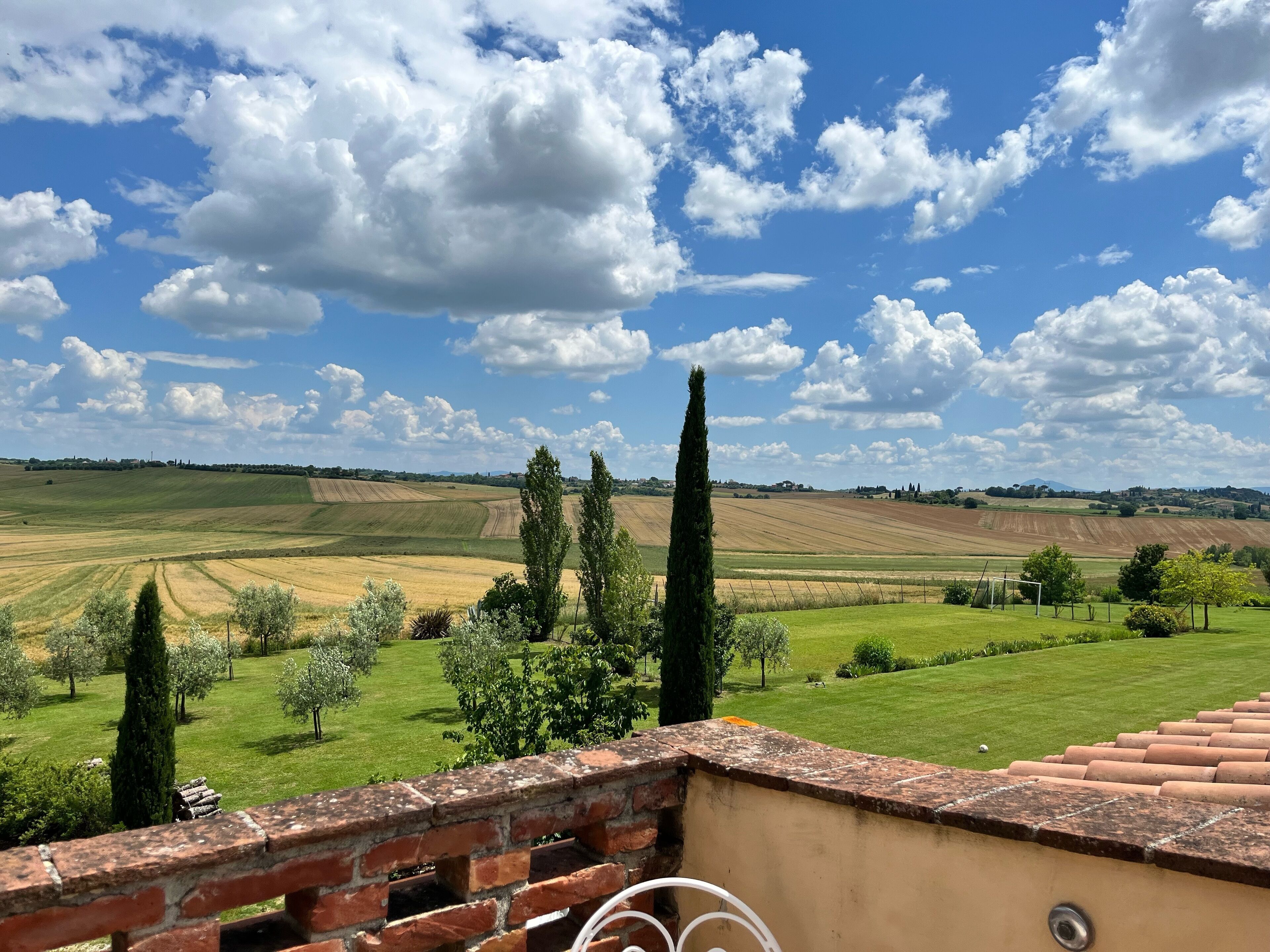 A view from a balcony with a brick wall, overlooking a rural landscape with fields and trees under a partly cloudy sky.