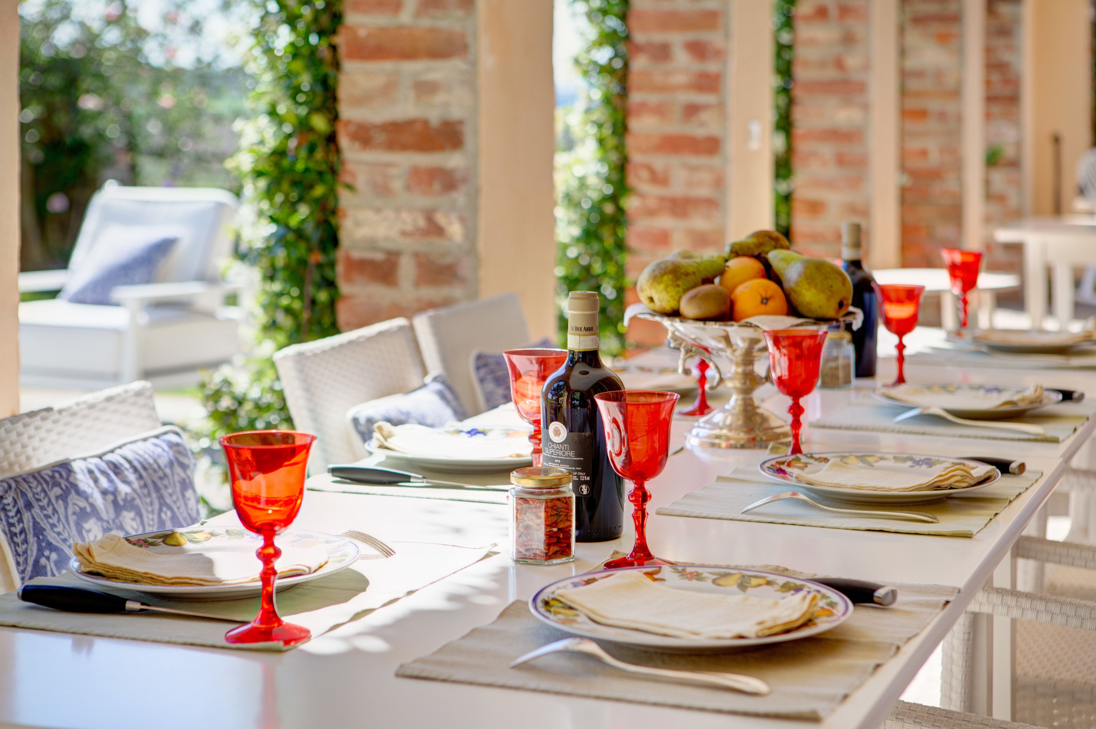 A dining table set with plates, cutlery, and red wine glasses, with a bottle of wine and a fruit bowl in the background.