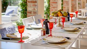 A dining table set with plates, cutlery, and red wine glasses, with a bottle of wine and a fruit bowl in the background.