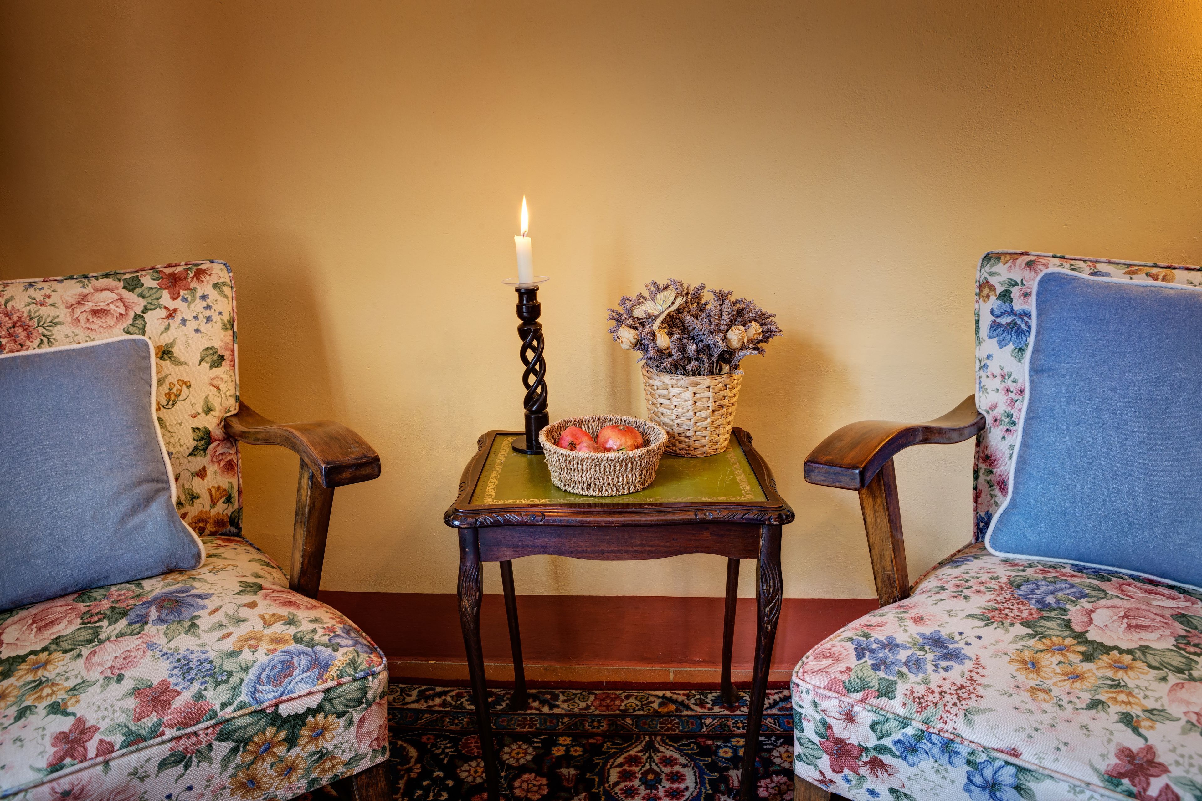 A room with two floral-patterned chairs, a small table with a candle, a basket of dried flowers, and a woven basket with apples.