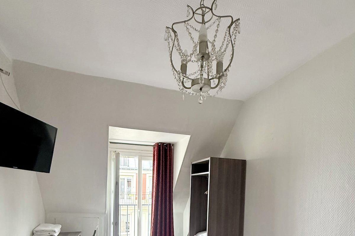 A hotel room with a bed, a desk, a chair, a wardrobe, a television, and a chandelier.