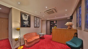 A hotel lobby with a red patterned carpet, a tufted orange sofa, a teal chair, a wooden reception desk, and a wall-mounted air conditioner. - Hôtel Louvre Richelieu (Paris)