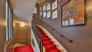 A staircase with red carpeted steps and a black railing, leading to a landing with framed artwork and a large colorful painting. - Hôtel Louvre Richelieu (Paris)