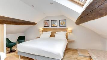 A bedroom with a sloped ceiling, a bed with white bedding, two green chairs, and artwork on the wall.