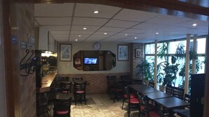 Interior de restaurante con suelo de patrón hexagonal, mesas y sillas de madera, un televisor montado en la pared y ventanas amplias con plantas afuera.