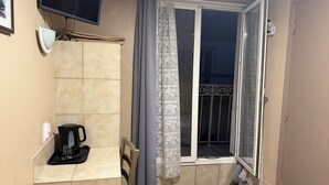 Una habitación pequeña y funcional con piso de baldosas, una ventana con cortina, televisor montado, cafetera y una silla.