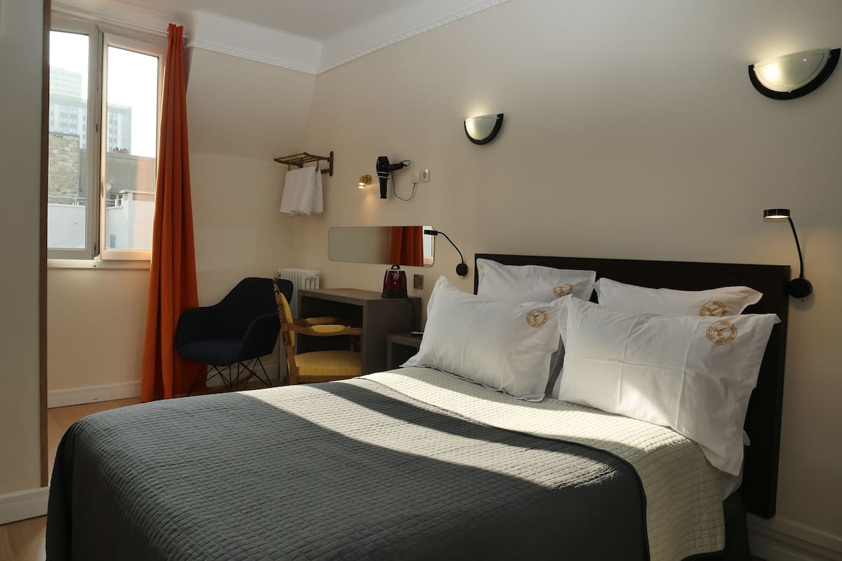 A hotel room with a bed, a desk, a chair, a window with curtains, and wall-mounted lighting.