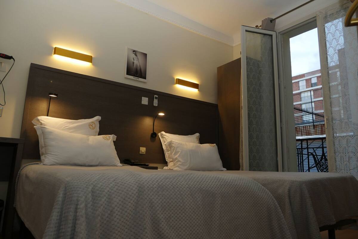 A hotel room with a bed, two bedside lamps, a telephone, and a window with a view of buildings.