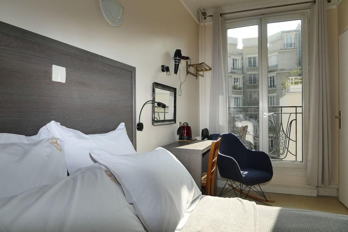 A hotel room with a bed, a desk, a chair, a mirror, and a view of the outside.