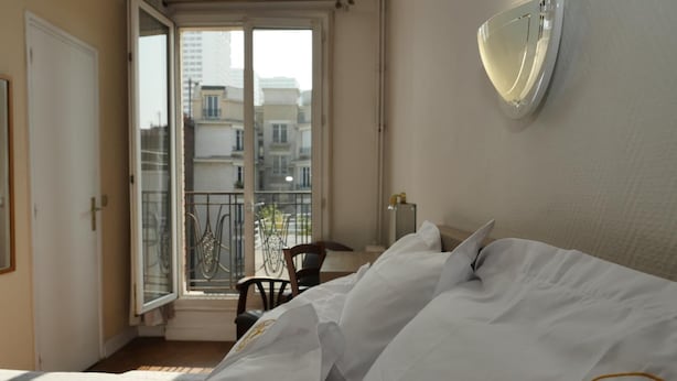 Quarto de hotel com cama, janela com vista para edifícios e varanda com uma cadeira.