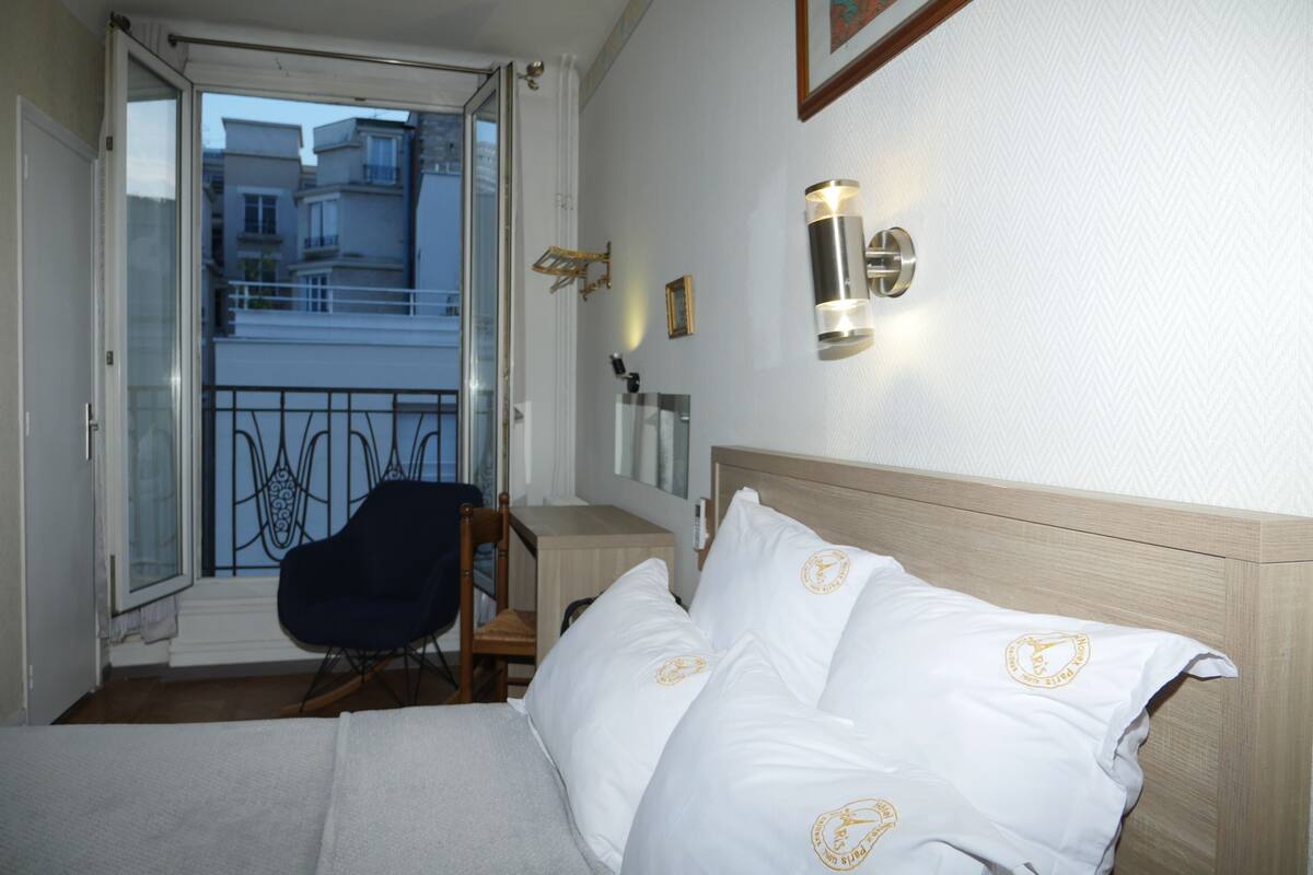 A hotel room with a bed, a chair, a desk, and a balcony with a view of buildings.