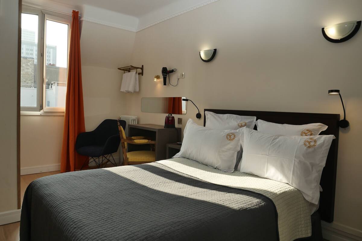 A hotel room with a large bed, a desk, a chair, a window with curtains, and wall-mounted lights.