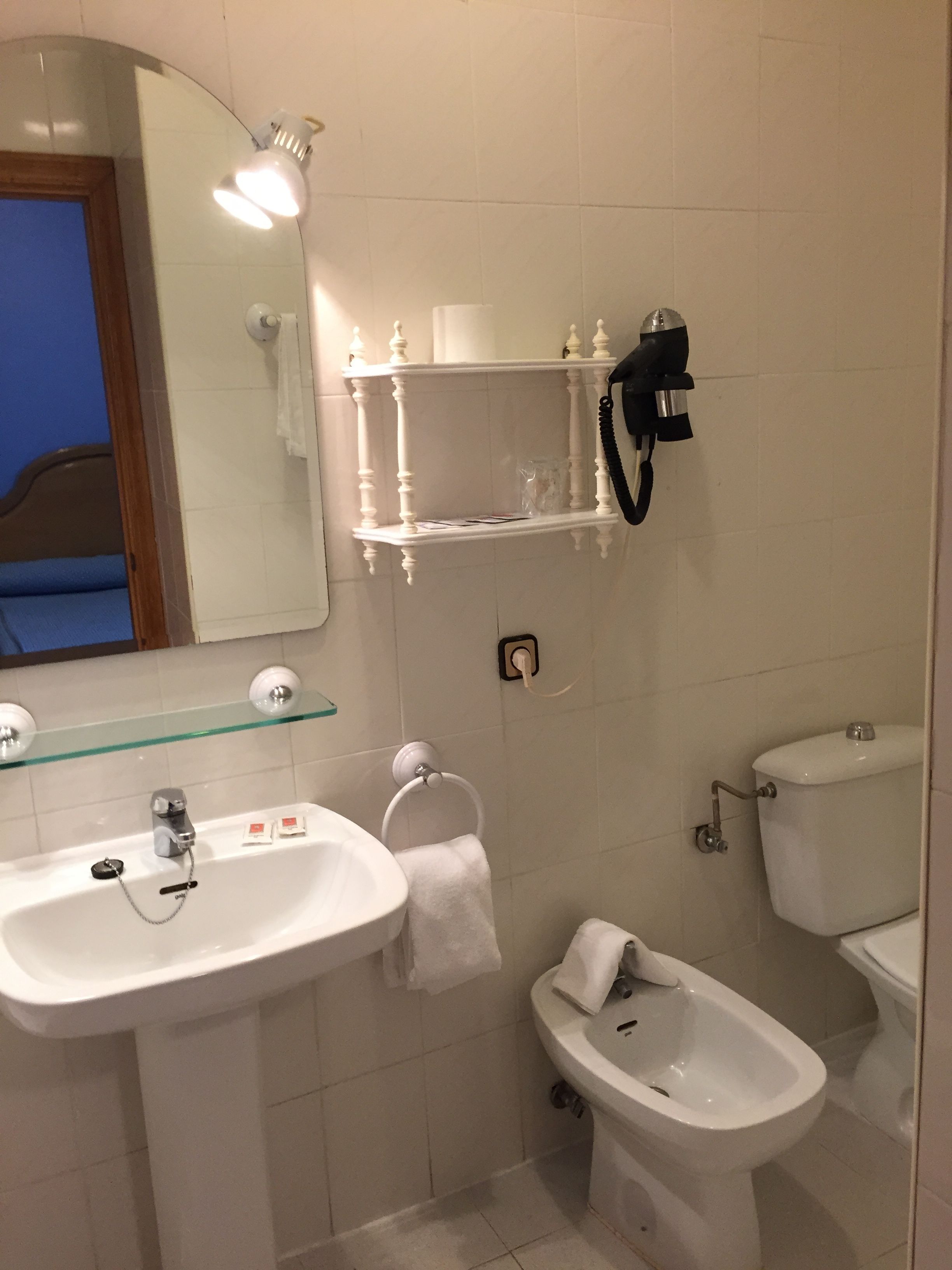 Un baño con lavamanos blanco, repisa de vidrio sobre el espejo, inodoro y bidé.