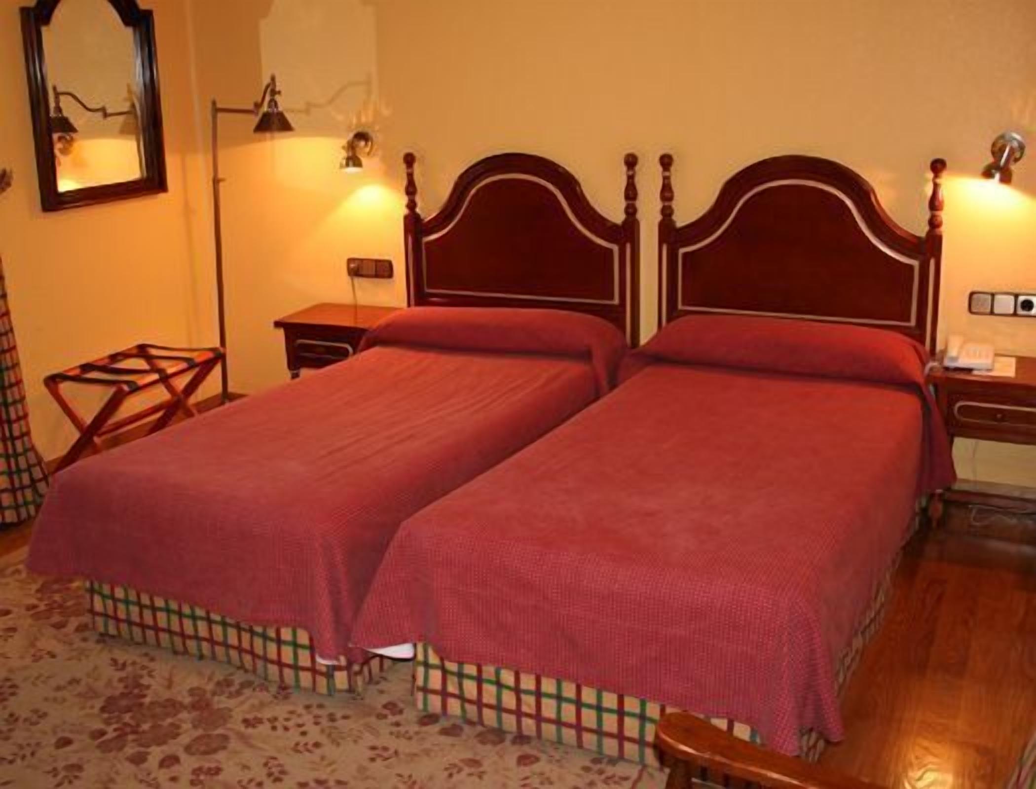 A room with two single beds, a nightstand with a lamp, a chair, and a patterned rug.
