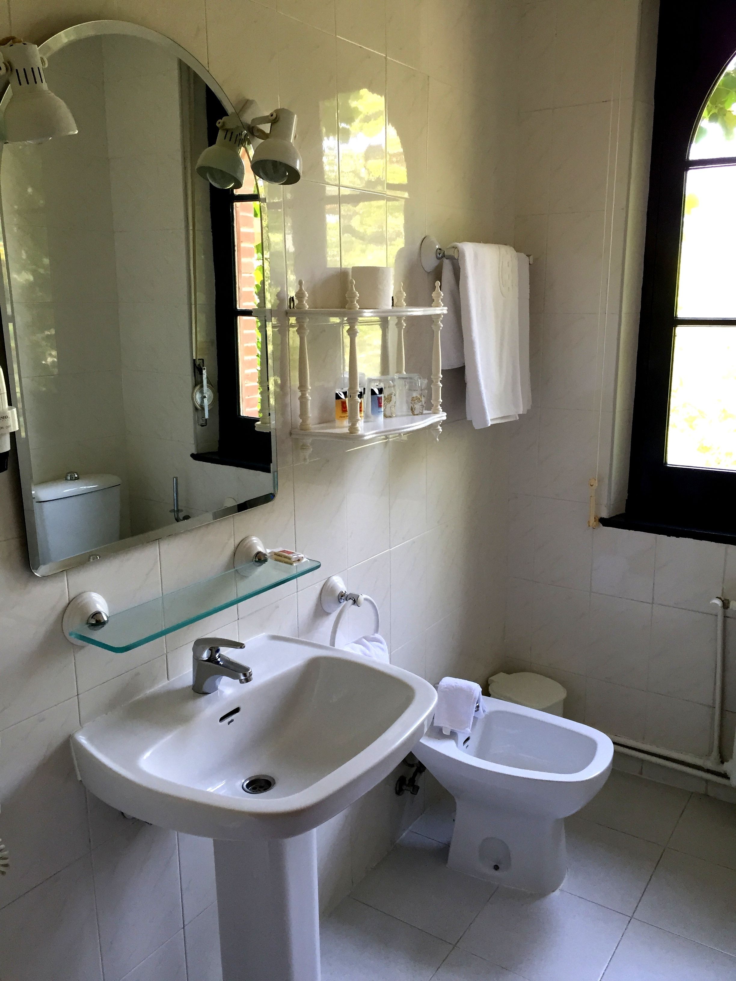Et badeværelse med en hvid vask, en bidet, en glashylde med toiletartikler, et spejl og et vindue med naturligt lys.