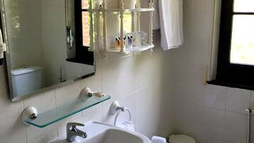 Et badeværelse med en hvid vask, en bidet, en glashylde med toiletartikler, et spejl og et vindue med naturligt lys.