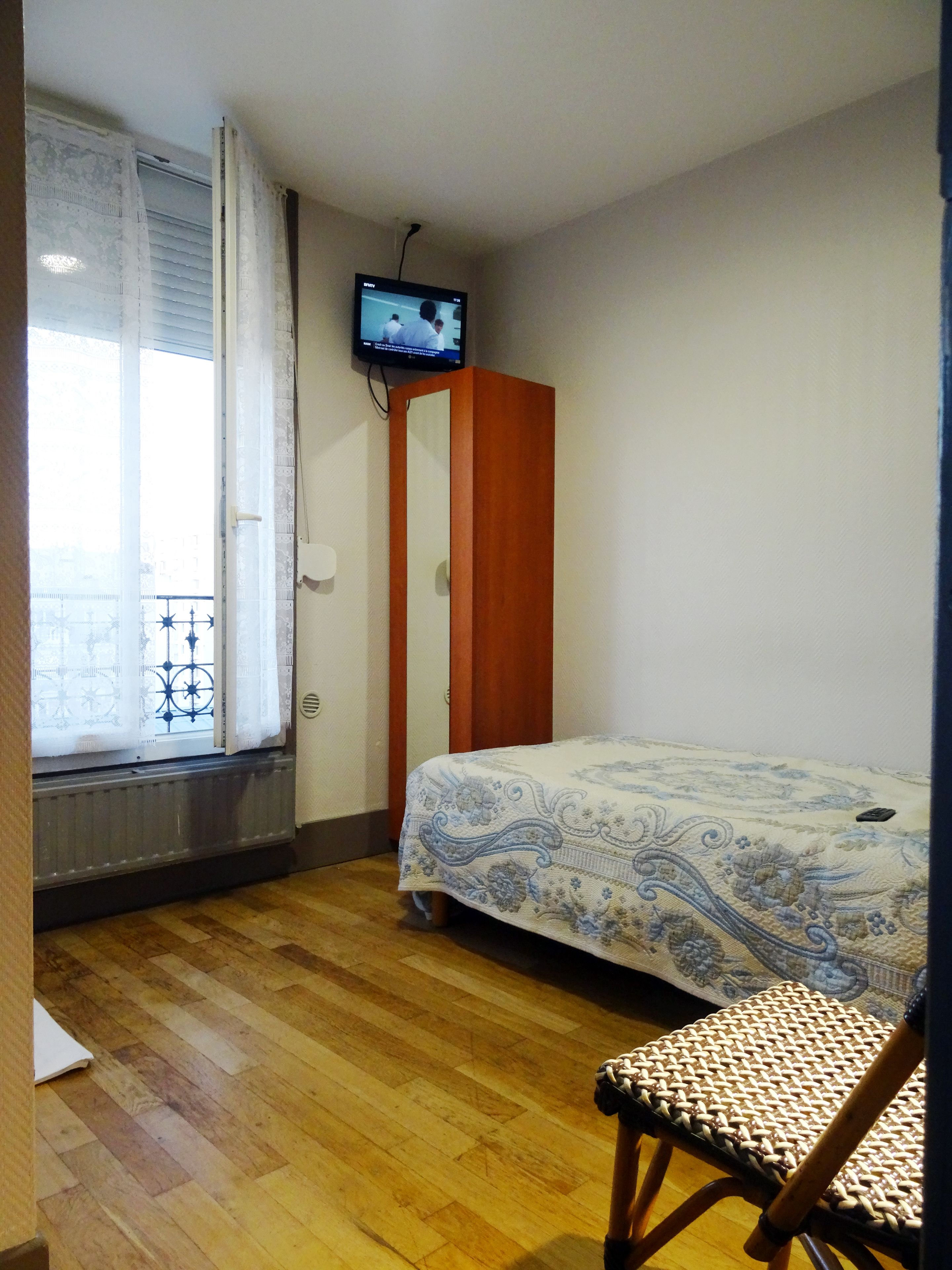Une petite pièce propre et bien rangée, avec un plancher en bois, un lit recouvert d’une couvre-lit à motifs, une armoire en bois, un téléviseur fixé au mur et une fenêtre donnant sur un balcon.