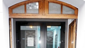 Uma entrada com portas duplas, moldura de madeira e painéis de vidro, que dá acesso a um corredor revestido de azulejos.