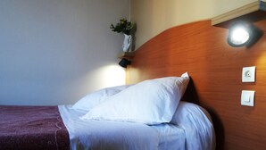 Uma cama bem arrumada com lençóis brancos, cabeceira de madeira e uma pequena mesa de cabeceira com luminária.