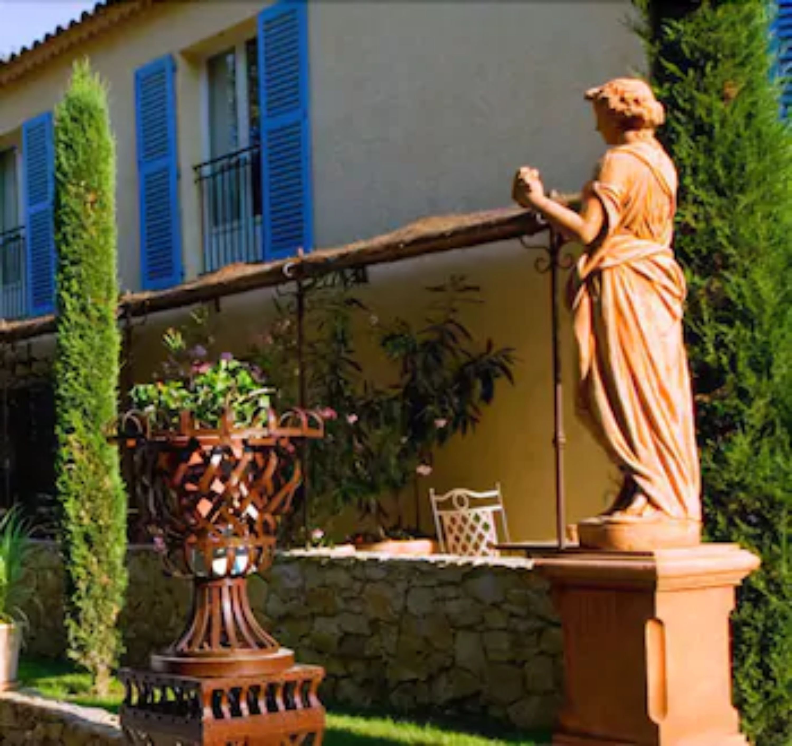 A statue in a garden with a stone wall and a building with blue shutters in the background.