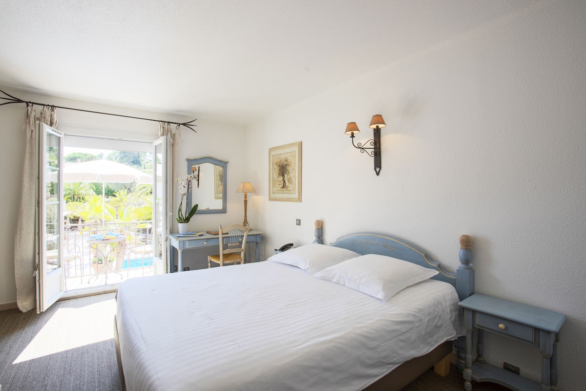 A bedroom with a bed, a desk, a chair, a nightstand, and a wall-mounted lamp.