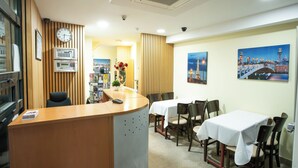 Un vestíbulo de hotel con mostrador de recepción, varias mesas de comedor y sillas. Hay relojes y una cámara de seguridad en la pared.