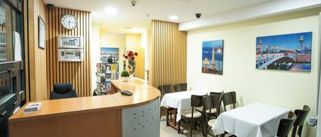 A hotel lobby with a reception desk, several dining tables, and chairs. There are clocks and a security camera on the wall.