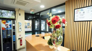 Un mostrador de recepción con un florero de flores rojas, un extintor, una máquina expendedora y un cartel con información del hotel.