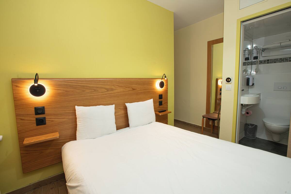 Une chambre d’hôtel avec une tête de lit en bois, deux tables de chevet, un lit avec du linge de maison blanc et une salle de bain visible à travers une porte ouverte.