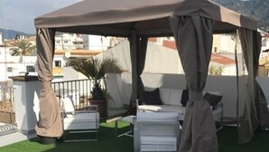 A rooftop terrace with a covered seating area, artificial grass, and a view of the cityscape and mountains. - Hotel Central (Marbella)