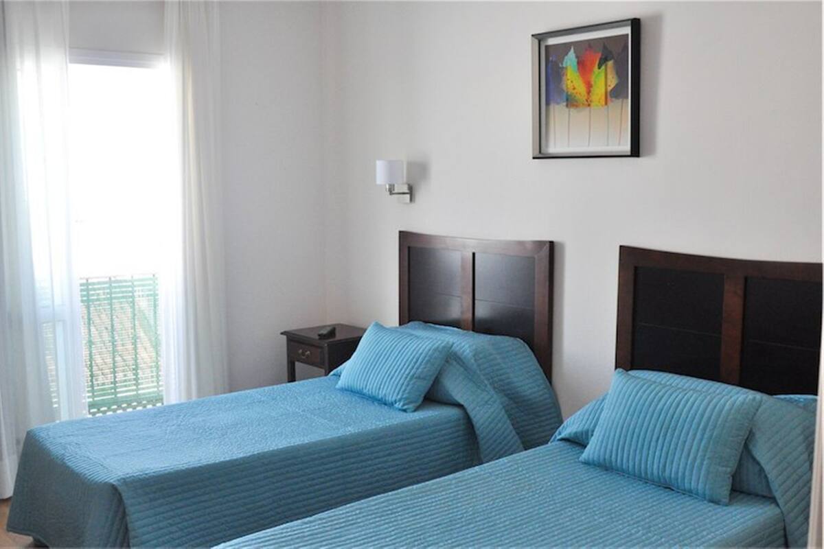 Quarto de hotel com duas camas, uma mesa de cabeceira, uma luminária na parede e uma janela com cortinas.