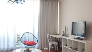 A modern living room with a round white ottoman, a black chair with a red cushion, a dining table, a television on a white shelf, and a large window with sheer curtains. - Hotel Central (Marbella)