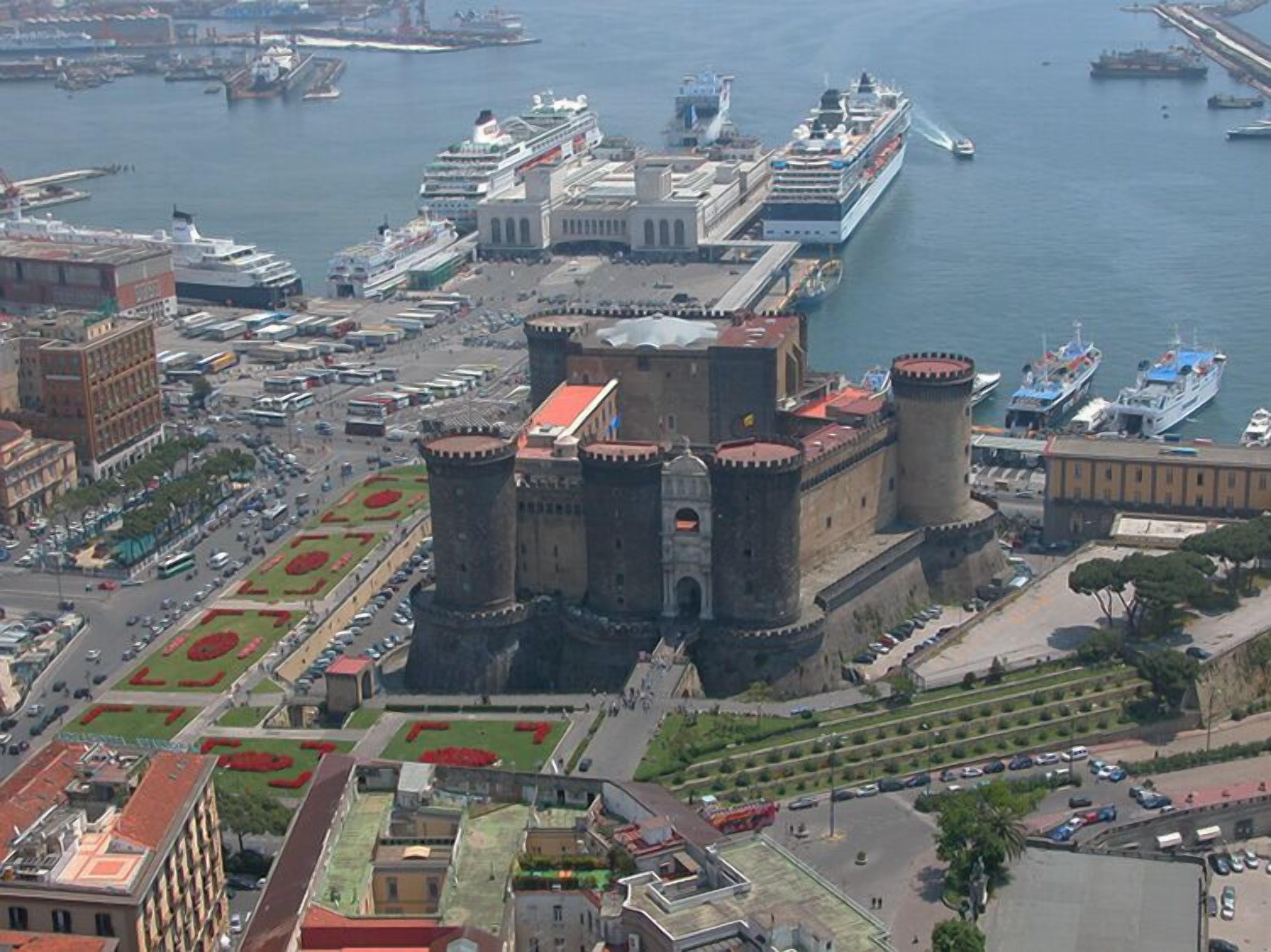 Un castello storico con struttura fortificata, circondato da edifici moderni e un porto turistico con navi da crociera.