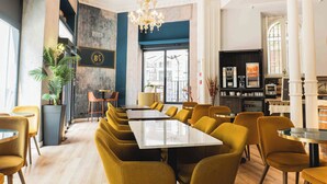 Een restaurant met een lange tafel, gele stoelen, een kroonluchter en uitzicht op de straat door een groot raam.