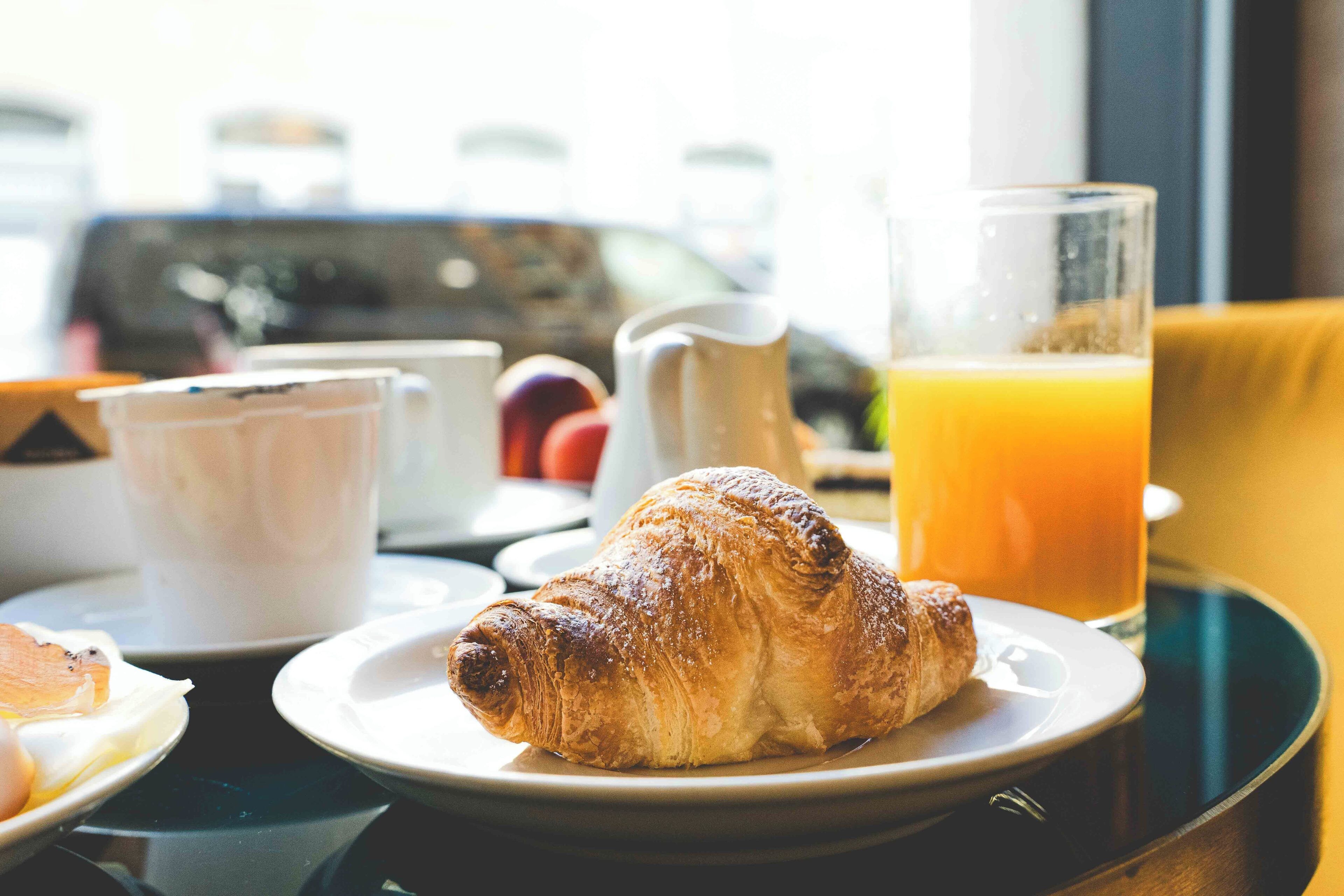 Un desayuno con un croissant, un vaso de jugo de naranja y una jarrón sobre una bandeja.