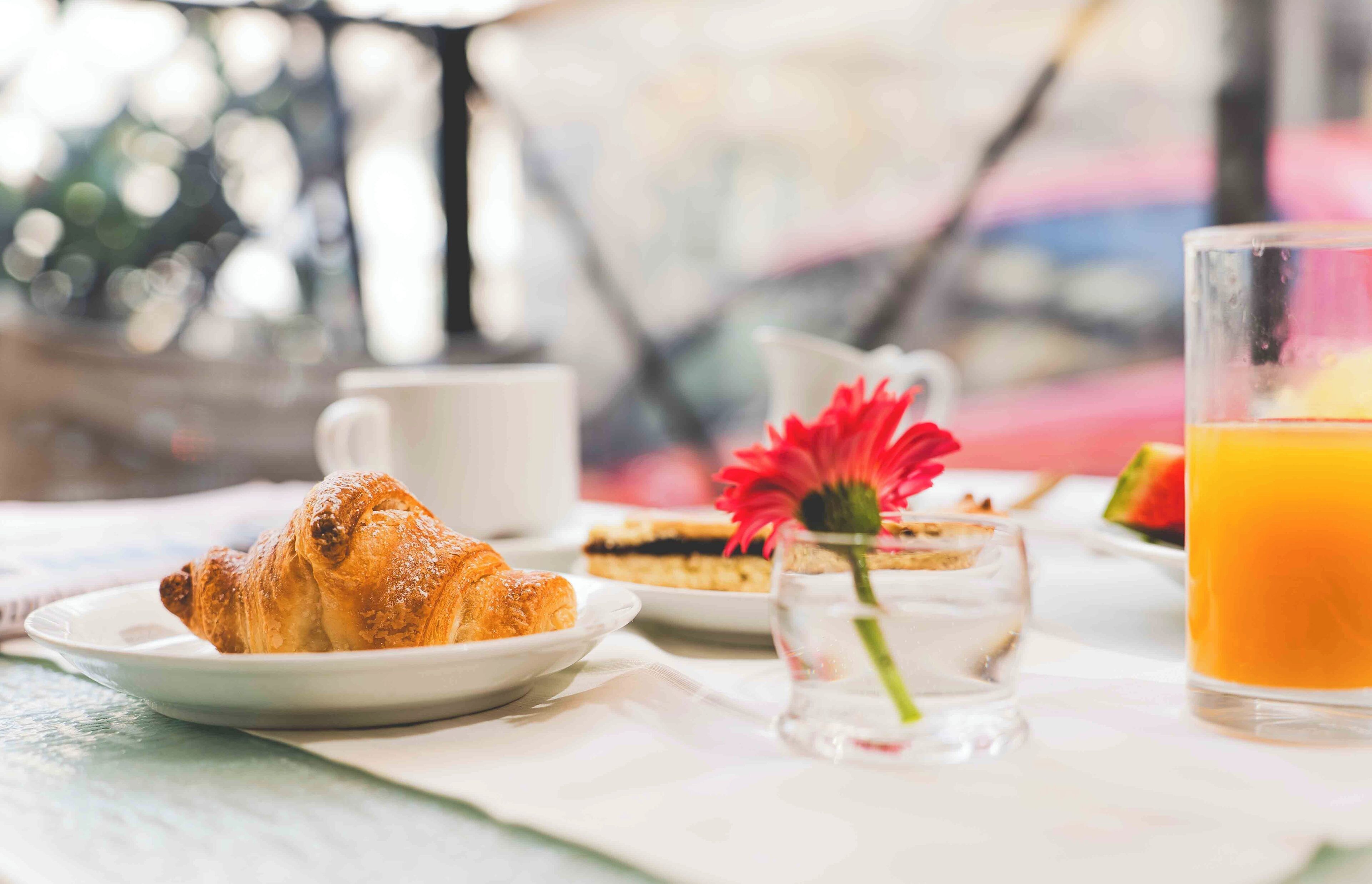 A breakfast setting with croissants, a glass of orange juice, a small vase with a flower, and a plate with a dessert.