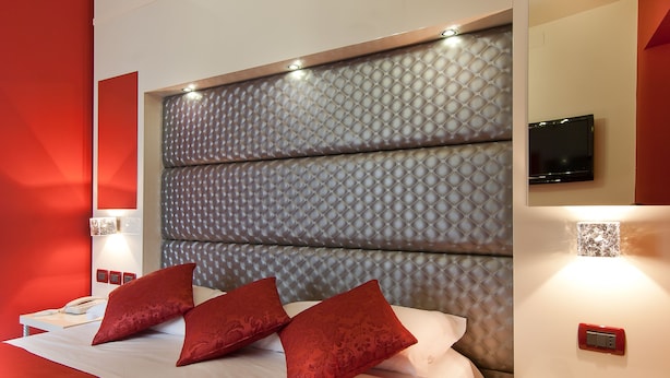 Een moderne hotelkamer met een groot bed, rode kussens, een wandtelevisie en een nachtkastje met afstandsbediening.