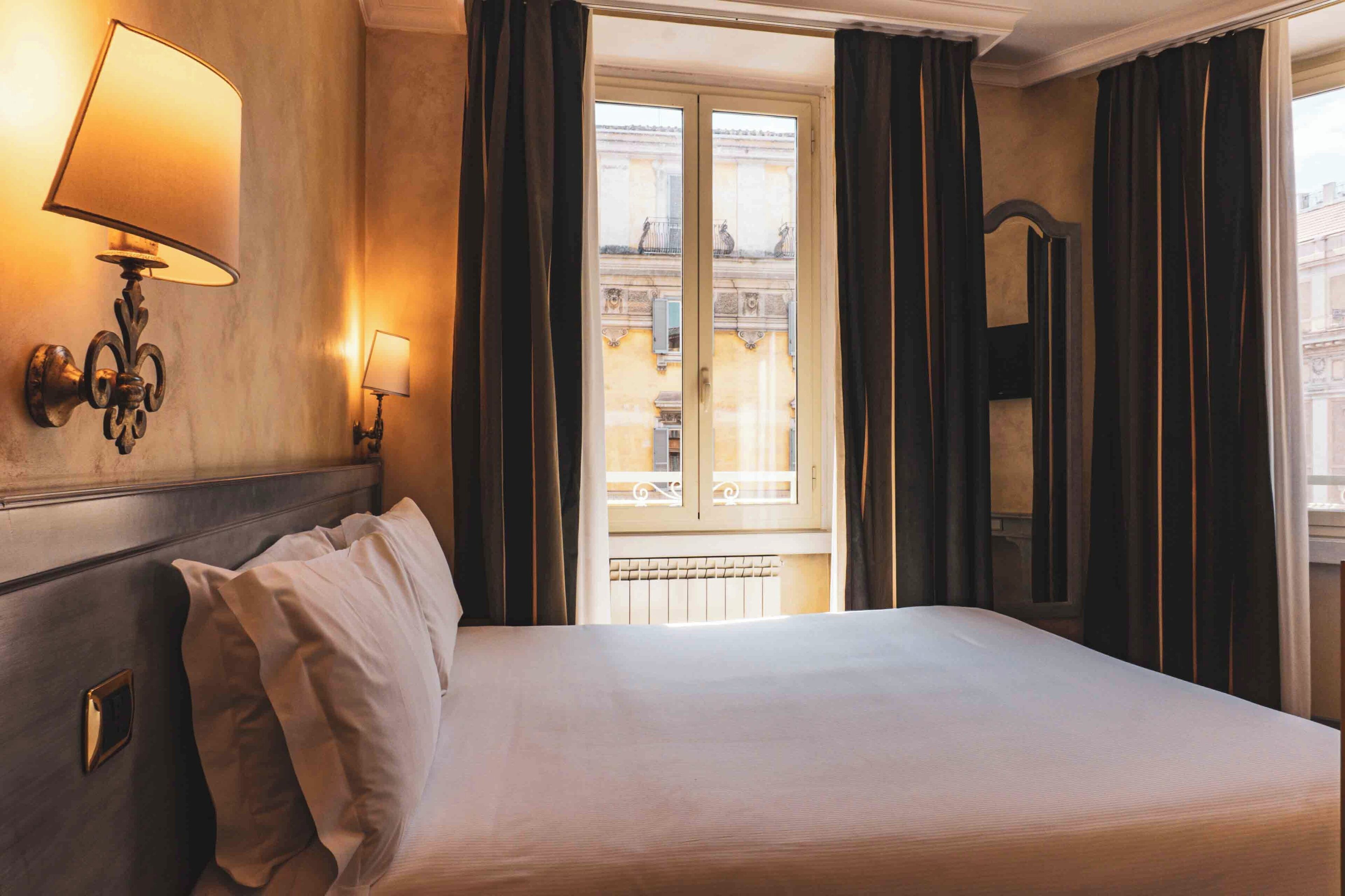 A hotel room with a large bed, two wall-mounted lamps, a window with curtains, and a view of buildings outside.