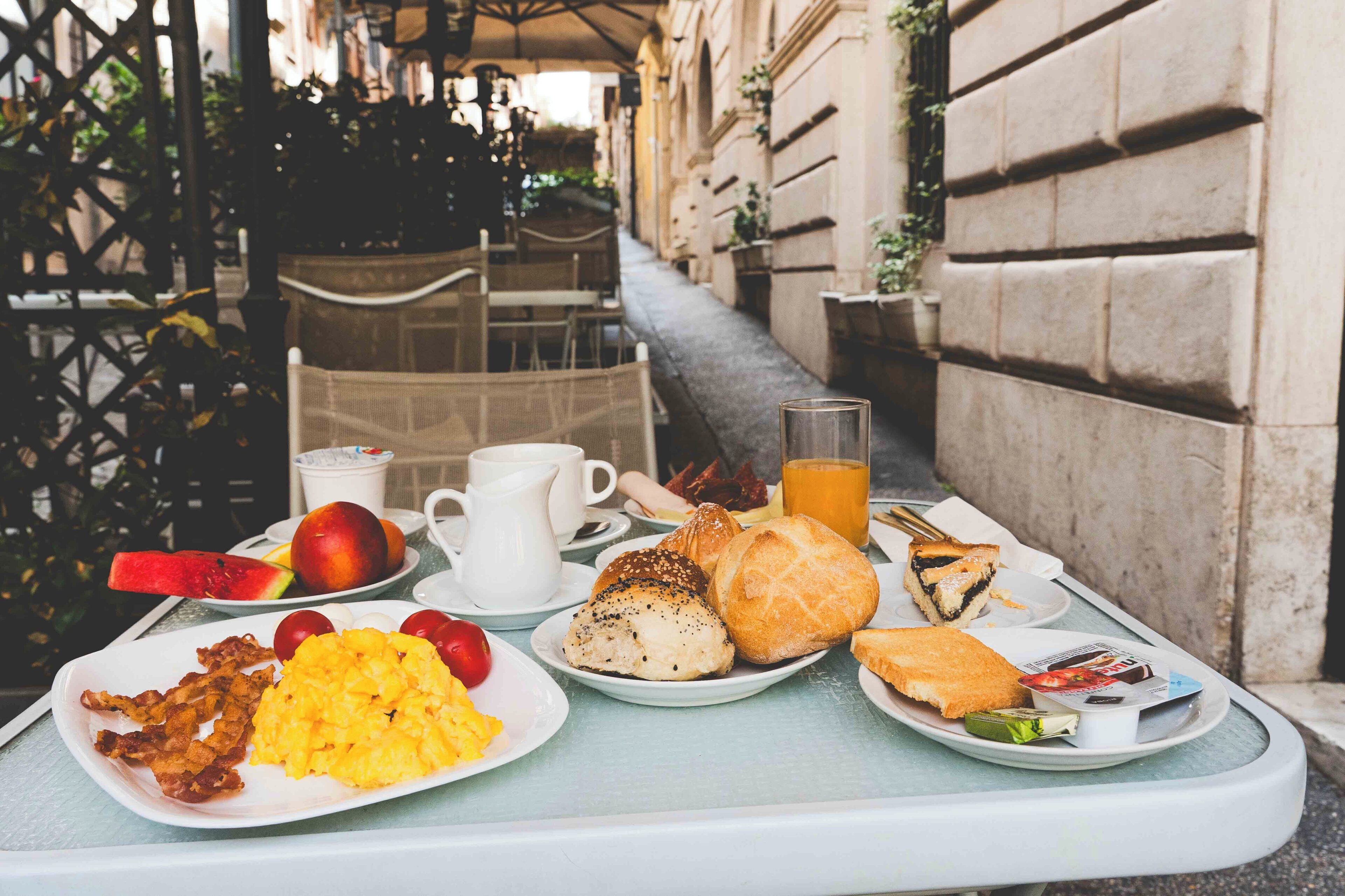 Un vassoio per la colazione con uova, pancetta, pane tostato, frutta e succo su un tavolo all'esterno di un edificio.