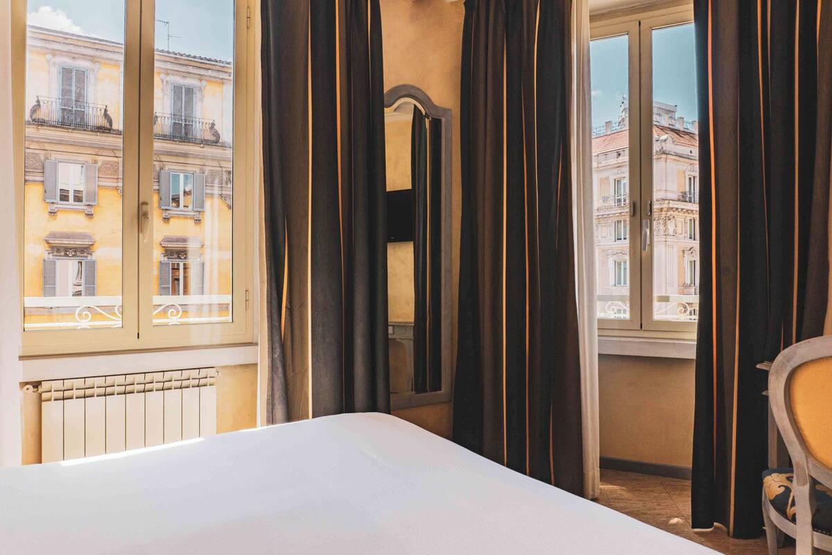 Ein Hotelzimmer mit Bett, Sessel, Fenster mit Vorhängen und Blick auf Gebäude.