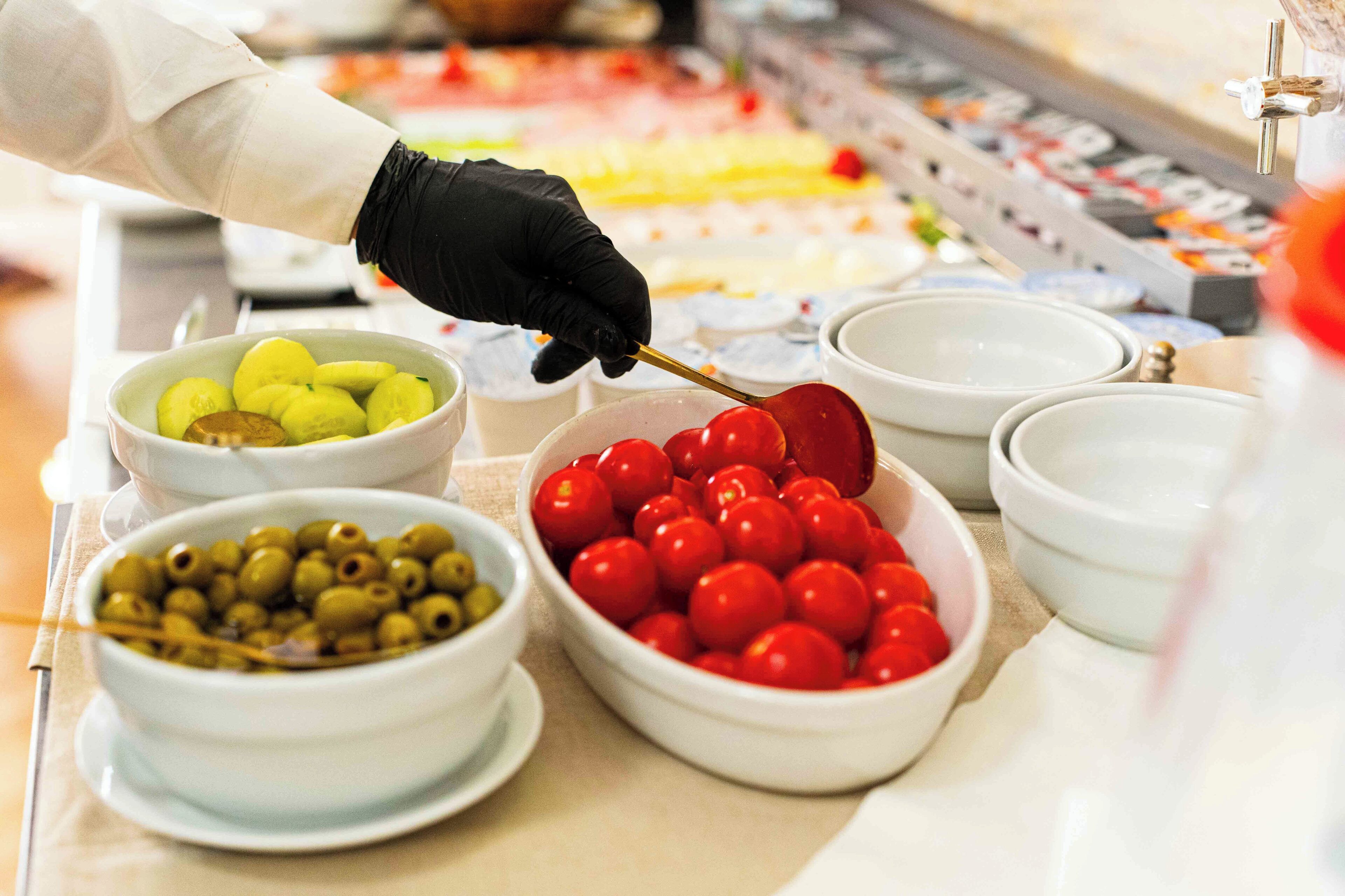 A person in a white coat and black gloves preparing food, with bowls of olives and cherry tomatoes visible.