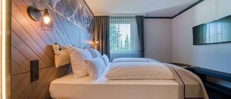 Una habitación de hotel moderna con una cama grande, cabecero de madera y vistas a árboles por la ventana.